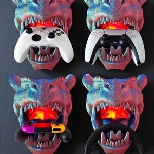 May include: Four dinosaur head-shaped wall mounts in blue, red, and purple hues, each holding a different gaming accessory: a white Xbox controller, a white and black PlayStation controller, a Nintendo Switch, and a black steering wheel with headphones.