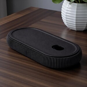 K&ouml;nnte beinhalten: Eine schwarze, ovale Dekoschale mit geripptem Rand und einer kleinen kreisf&ouml;rmigen &Ouml;ffnung. Die Schale besteht aus einem matten Material und liegt auf einer dunklen Holzoberfl&auml;che. Eine wei&szlig;e, geometrische Vase mit einer gr&uuml;nen Pflanze befindet sich im Hintergrund.