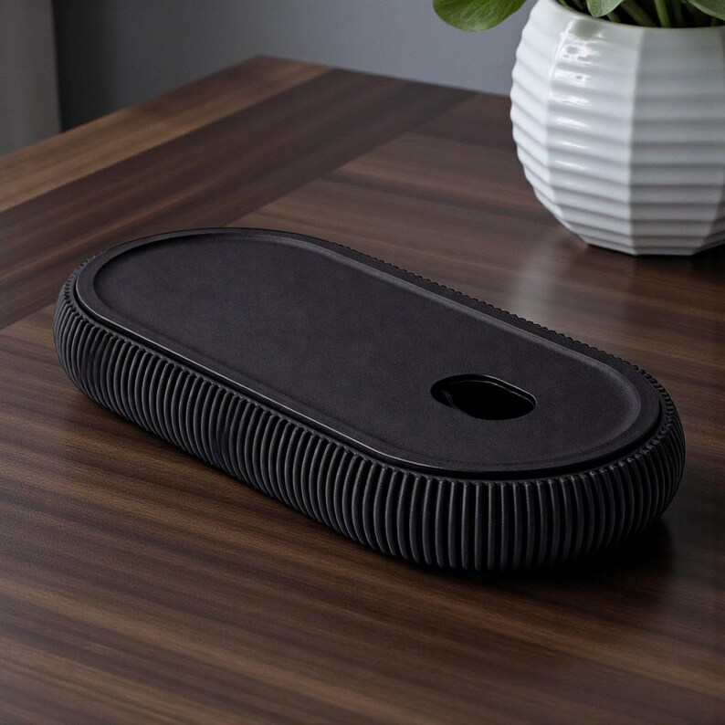 K&ouml;nnte beinhalten: Eine schwarze, ovale Dekoschale mit geripptem Rand und einer kleinen kreisf&ouml;rmigen &Ouml;ffnung. Die Schale besteht aus einem matten Material und liegt auf einer dunklen Holzoberfl&auml;che. Eine wei&szlig;e, geometrische Vase mit einer gr&uuml;nen Pflanze befindet sich im Hintergrund.