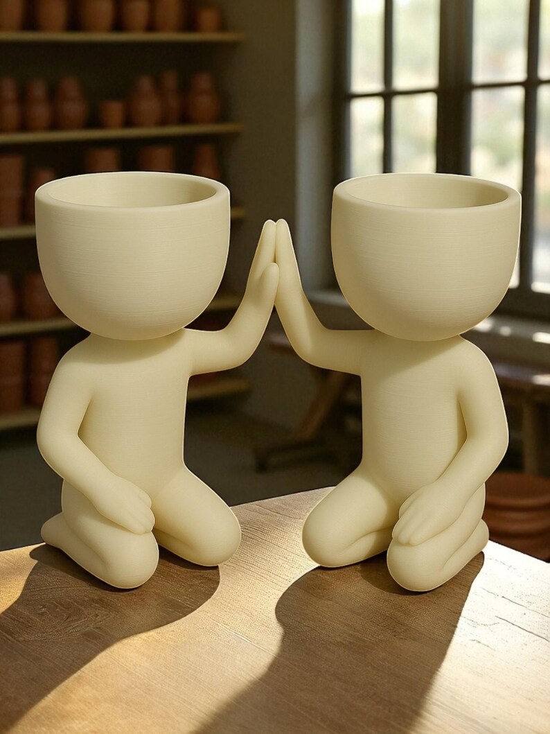May include: Two cream-colored, kneeling human-shaped planters with open bowl-shaped heads. The figures are positioned with their hands raised in a high-five gesture. The planters are on a wooden surface, with a blurred background of shelves and windows.