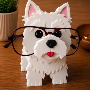May include: A white dog-shaped eyeglasses holder with a black nose, red tongue, and pink inner ears. The holder is wearing a pair of dark-rimmed glasses. The holder is made of a textured material and sits on a wooden surface.