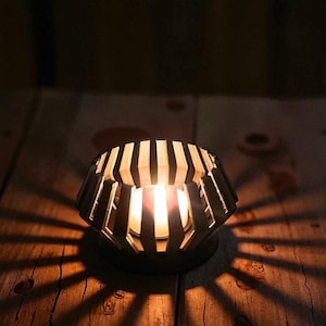 Puede incluir: Un portavelas decorativo con dise&ntilde;o geom&eacute;trico. El soporte marr&oacute;n oscuro tiene ranuras verticales que proyectan patrones de luz y sombra sobre la superficie de madera. Una vela encendida crea un ambiente c&aacute;lido.
