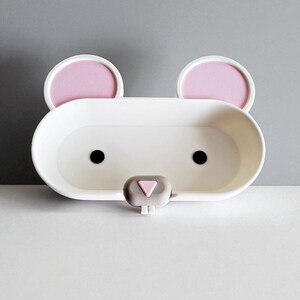 May include: A white, bear-shaped soap dish with pink ear accents and black eyes. The dish has a small, gray nose and is designed to hold soap or other small items. The overall design is simple and playful, suitable for a bathroom or children's room.