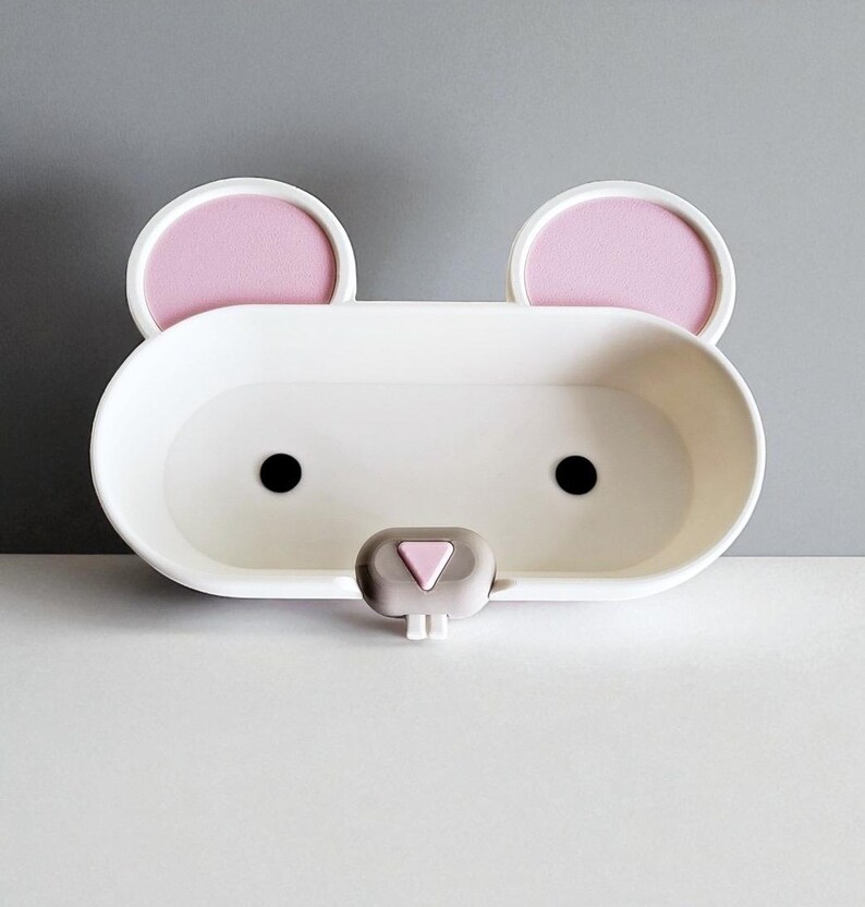 May include: A white, bear-shaped soap dish with pink ear accents and black eyes. The dish has a small, gray nose and is designed to hold soap or other small items. The overall design is simple and playful, suitable for a bathroom or children's room.