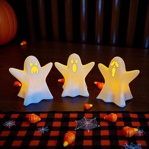 May include: Three white ghost-shaped Halloween decorations with glowing yellow faces and outstretched arms. The ghosts are on a dark wooden surface, surrounded by candy corn and small pumpkin decorations. A black and red checkered fabric is in the foreground.