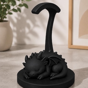May include: A black dragon figurine resting on a round base, with a curved support extending upwards. The dragon has closed eyes, horns, and wings. The figurine is a decorative item.