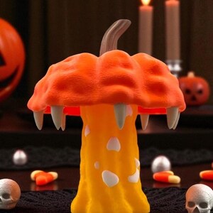 May include: A Halloween-themed decorative mushroom with an orange cap and yellow stem. The cap features fangs and the stem has white spots. The background includes candles, pumpkins, and skull decorations, creating a festive atmosphere.