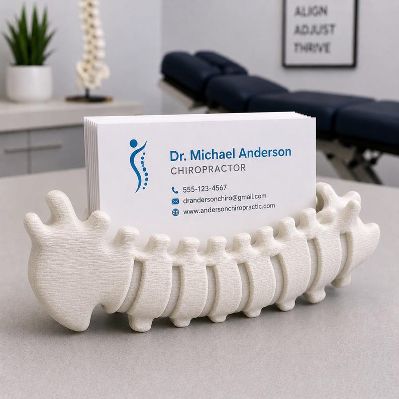 Op de afbeelding: Een cr&egrave;mekleurige, ruggengraatvormige visitekaarthouder met een stapel witte visitekaartjes. De kaarten tonen "Dr. Michael Anderson, Chiropractor" en contactgegevens. De achtergrond toont een chiropractietafel en een bord met "Align, Adjust, Thrive".