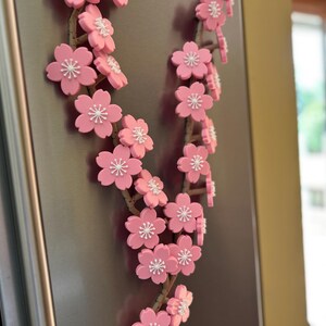 May include: A decorative floral garland featuring pink cherry blossoms with white centers. The blossoms are attached to a brown, twig-like stem. The garland is displayed on a metallic surface.