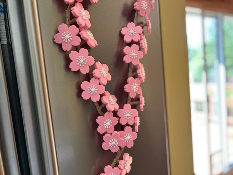May include: A decorative floral garland featuring pink cherry blossoms with white centers. The blossoms are attached to a brown, twig-like stem. The garland is displayed on a metallic surface.