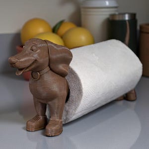 May include: A brown dachshund-shaped paper towel holder. The dog has a collar and shoes, and the paper towel roll is inserted through its body. The holder is on a gray countertop, with lemons and other kitchen items in the background.