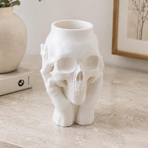 May include: A white ceramic skull-shaped vase with a wide opening. The skull is held by two hands, creating a unique decorative piece. The vase is placed on a light-colored surface, suitable for home decor.