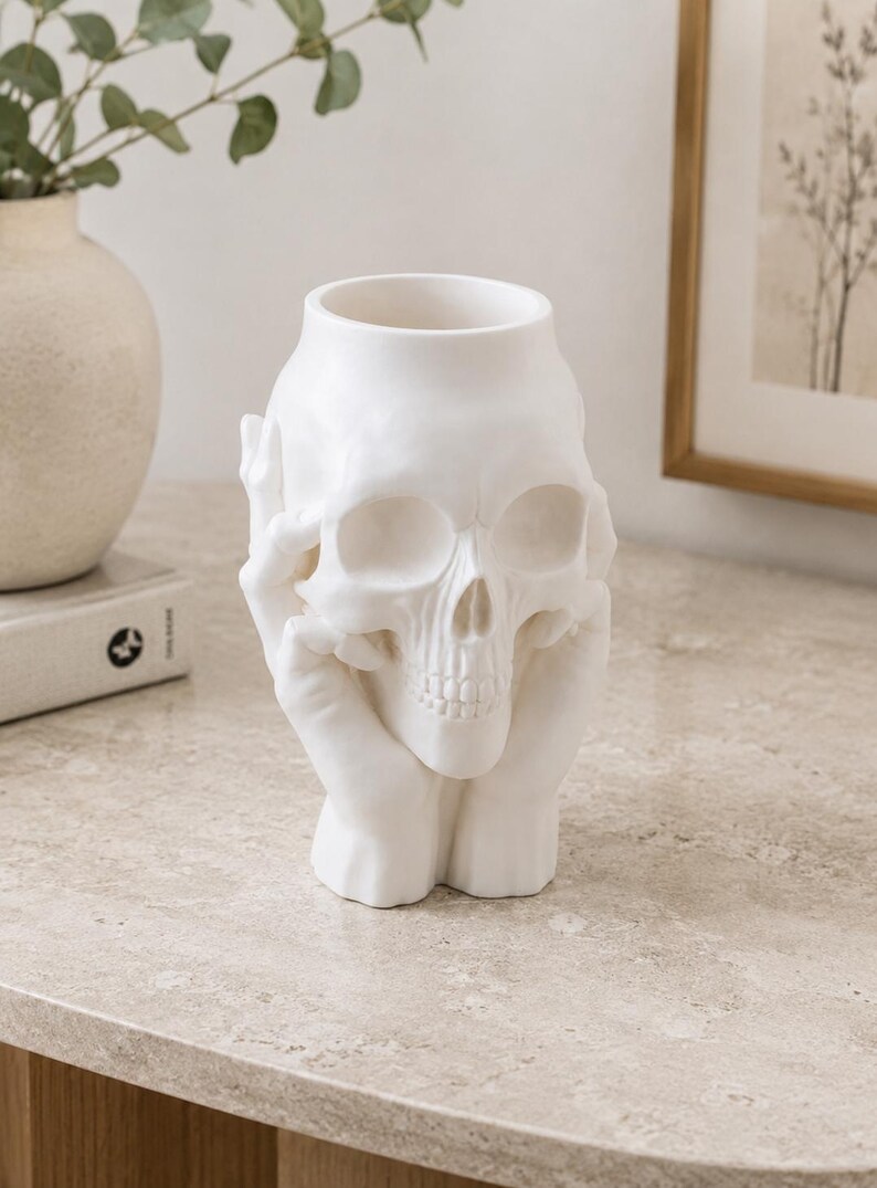 May include: A white ceramic skull-shaped vase with a wide opening. The skull is held by two hands, creating a unique decorative piece. The vase is placed on a light-colored surface, suitable for home decor.