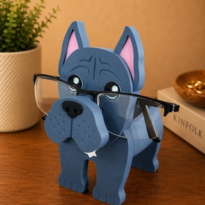 May include: A blue wooden bulldog-shaped glasses holder with pink inner ears and a black nose. The holder is designed to hold eyeglasses, with the glasses resting on the dog's nose. The dog is sitting on a wooden surface.