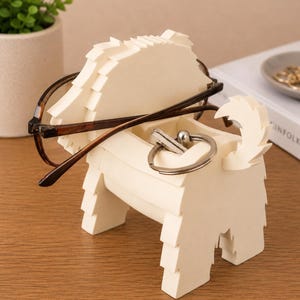 May include: A cream-colored dog-shaped desk organizer holding eyeglasses and a silver keychain. The organizer has a textured, layered design and is sitting on a wooden surface. A small potted plant is visible in the background.