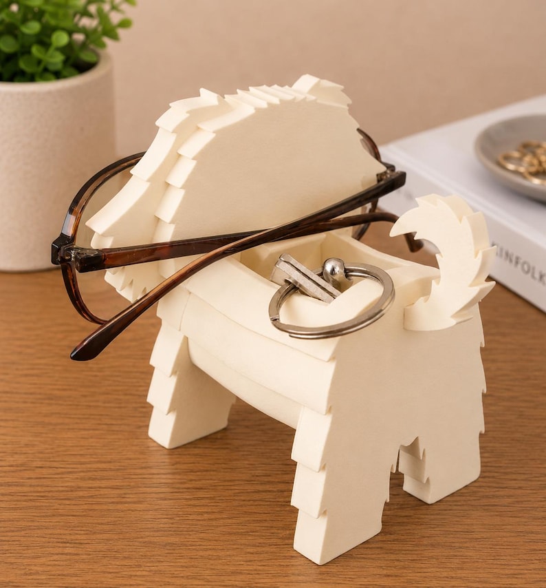 May include: A cream-colored dog-shaped desk organizer holding eyeglasses and a silver keychain. The organizer has a textured, layered design and is sitting on a wooden surface. A small potted plant is visible in the background.