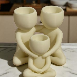 Puede incluir: Una escultura familiar de color crema. Dos figuras abrazan a una figura m&aacute;s peque&ntilde;a, cada una con una cabeza redondeada y una abertura en forma de cuenco. Las figuras est&aacute;n arrodilladas o sentadas, creando una sensaci&oacute;n de unidad y protecci&oacute;n.