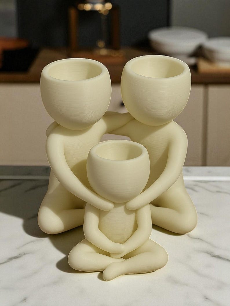 Puede incluir: Una escultura familiar de color crema. Dos figuras abrazan a una figura m&aacute;s peque&ntilde;a, cada una con una cabeza redondeada y una abertura en forma de cuenco. Las figuras est&aacute;n arrodilladas o sentadas, creando una sensaci&oacute;n de unidad y protecci&oacute;n.