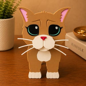 May include: A brown cat figurine with large, expressive eyes, pink-lined ears, and white whiskers. The cat has a red nose and a white muzzle. The figurine is made of layered materials and stands upright on a wooden surface.