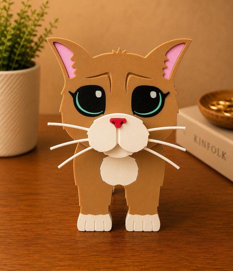 May include: A brown cat figurine with large, expressive eyes, pink-lined ears, and white whiskers. The cat has a red nose and a white muzzle. The figurine is made of layered materials and stands upright on a wooden surface.