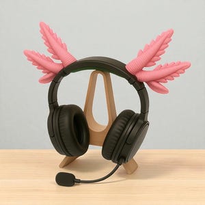 Puede incluir: Auriculares negros de diadema con micr&oacute;fono, adornados con decoraciones rosas inspiradas en el ajolote. Los auriculares se exhiben en un soporte de madera, mostrando un dise&ntilde;o &uacute;nico y divertido. El micr&oacute;fono est&aacute; conectado al auricular izquierdo.