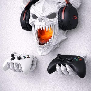May include: A white skull-shaped wall mount with a fiery interior, holding black and red headphones. Two game controllers, one white and one black, are held in skeletal hands below. A unique gaming accessory.