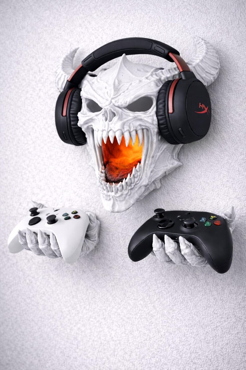 May include: A white skull-shaped wall mount with a fiery interior, holding black and red headphones. Two game controllers, one white and one black, are held in skeletal hands below. A unique gaming accessory.