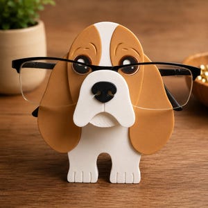 May include: A whimsical eyeglasses holder shaped like a beagle dog. The holder is primarily brown, white, and black, with the glasses resting on the dog's nose. The dog has large, expressive eyes and a friendly expression. A fun and functional desk accessory.