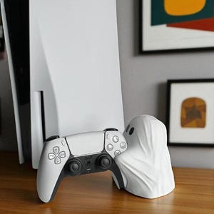 May include: A white PlayStation 5 console and controller are displayed with a white ghost-shaped accessory. The controller has black accents and buttons. The ghost accessory has two black eye holes. The items are on a wooden surface.