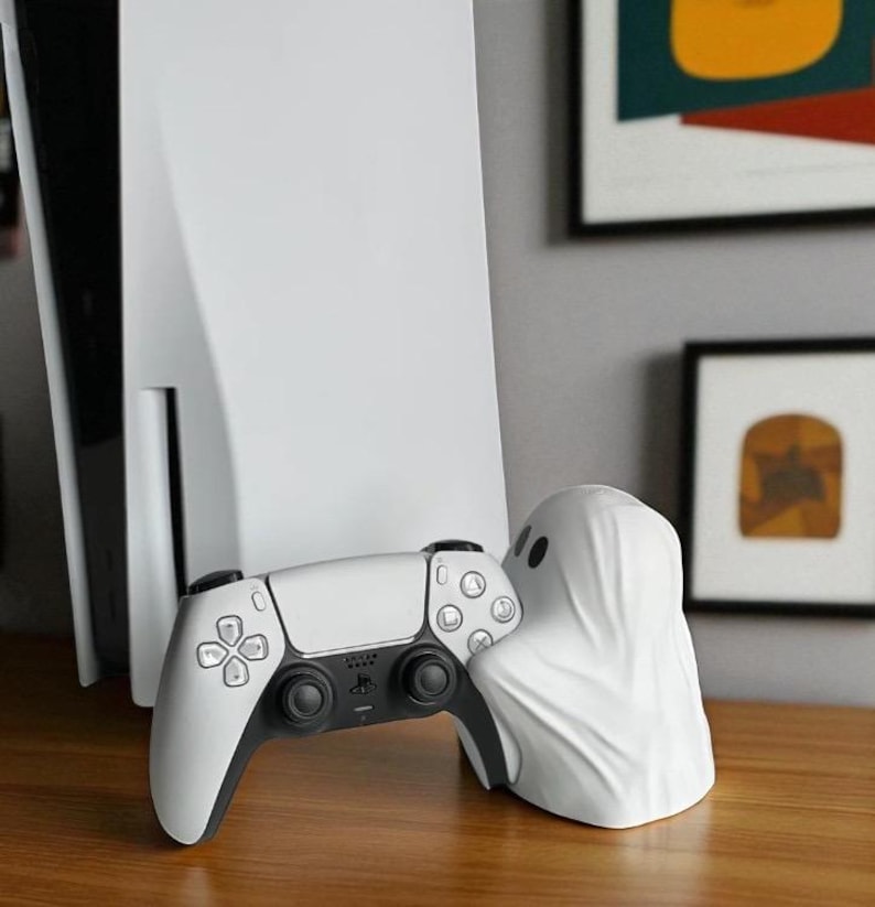 May include: A white PlayStation 5 console and controller are displayed with a white ghost-shaped accessory. The controller has black accents and buttons. The ghost accessory has two black eye holes. The items are on a wooden surface.