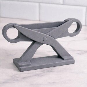 May include: A gray, scissor-shaped decorative stand. The stand features a rectangular base, angled legs, and two circular handles. The design is modern and minimalist, suitable for various home decor styles.