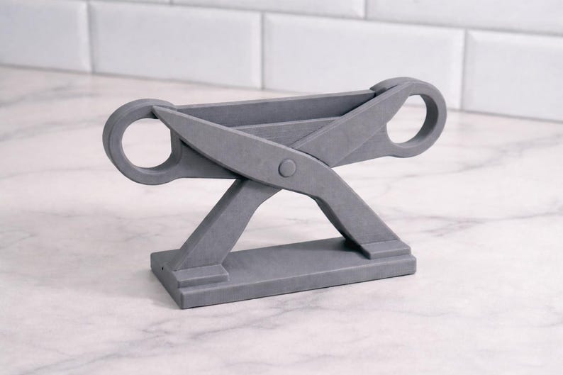 May include: A gray, scissor-shaped decorative stand. The stand features a rectangular base, angled legs, and two circular handles. The design is modern and minimalist, suitable for various home decor styles.