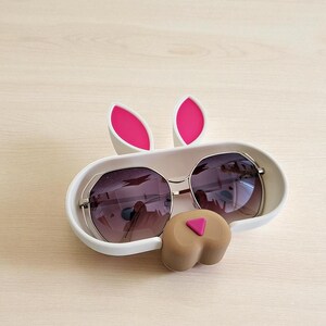 May include: A white bunny-shaped sunglasses holder with pink accents. The holder features two upright pink-lined ears and a brown nose with a pink triangle. Sunglasses with dark lenses rest inside the holder.