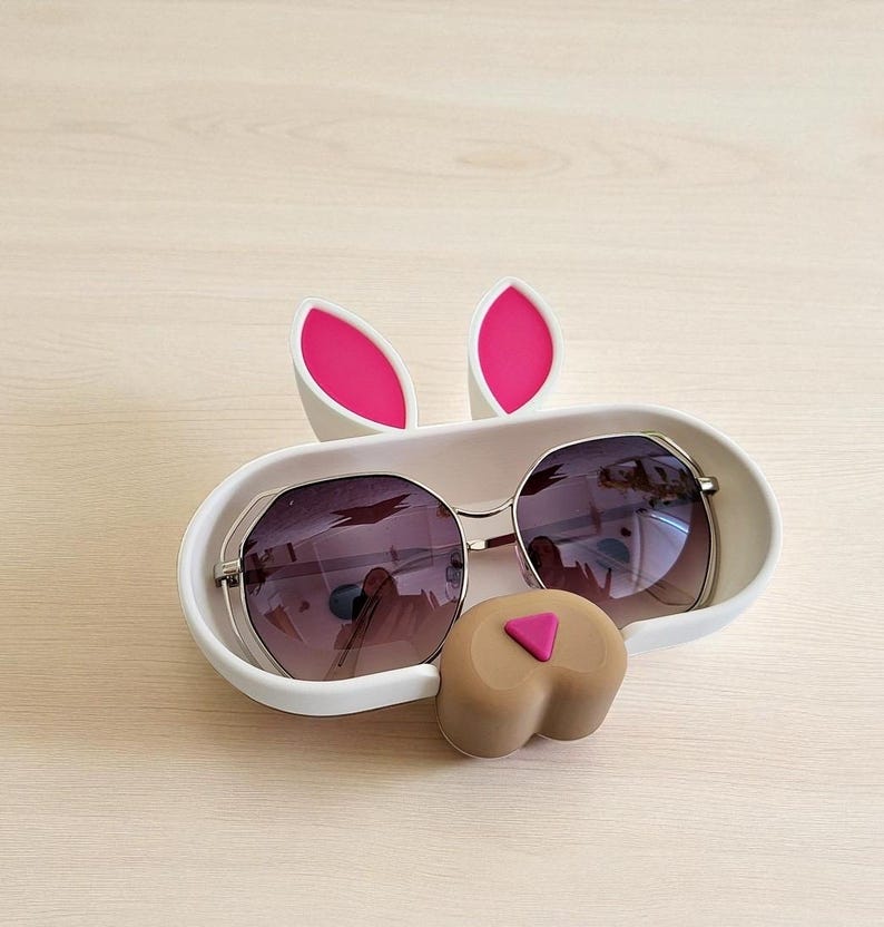 May include: A white bunny-shaped sunglasses holder with pink accents. The holder features two upright pink-lined ears and a brown nose with a pink triangle. Sunglasses with dark lenses rest inside the holder.