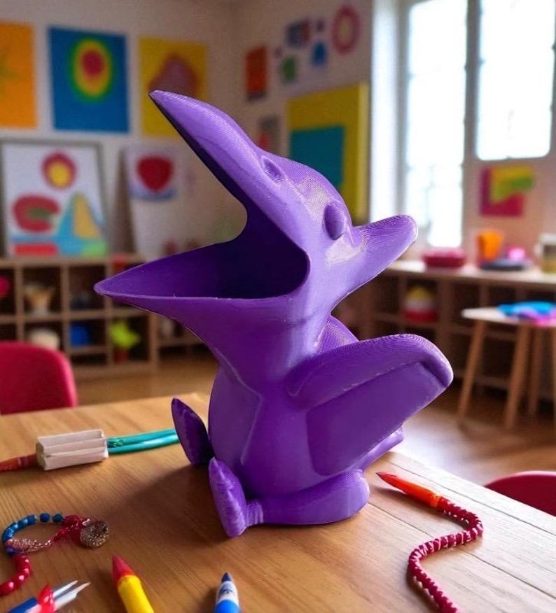 May include: A vibrant purple pterodactyl-shaped container, likely 3D-printed, with an open mouth and outstretched wings. The container sits on a wooden table, surrounded by colorful art supplies and a beaded bracelet. The design is whimsical and playful.