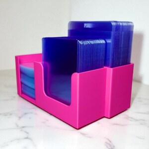 May include: A bright pink card holder with three compartments. The holder contains stacks of blue cards. The holder is made of a solid material and has a modern, minimalist design. The cards appear to be standard size.