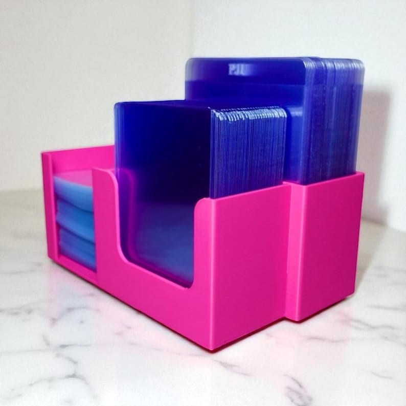 May include: A bright pink card holder with three compartments. The holder contains stacks of blue cards. The holder is made of a solid material and has a modern, minimalist design. The cards appear to be standard size.