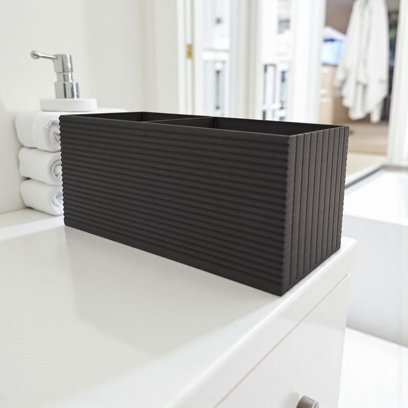May include: A dark gray rectangular bathroom organizer with a ribbed texture. The organizer has two compartments and is placed on a white surface. A white soap dispenser and rolled white towels are visible in the background.