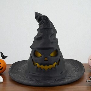May include: A black Halloween witch hat with a carved jack-o'-lantern face. The hat has a wide brim and a tall, pointed crown. The interior glows with a yellow light, creating a spooky effect. The hat is a decorative item.