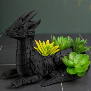 May include: A black dragon-shaped planter with a detailed, textured surface. The dragon holds several faux succulents in shades of green and yellow. The planter is sitting on a dark tiled surface.