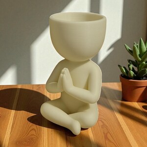 Puede incluir: Una figura crema, sentada con las manos juntas en oraci&oacute;n, dise&ntilde;ada como maceta. La cabeza es un cuenco grande y abierto para plantas. La figura est&aacute; sobre una superficie de madera, con una peque&ntilde;a planta suculenta en maceta al fondo.