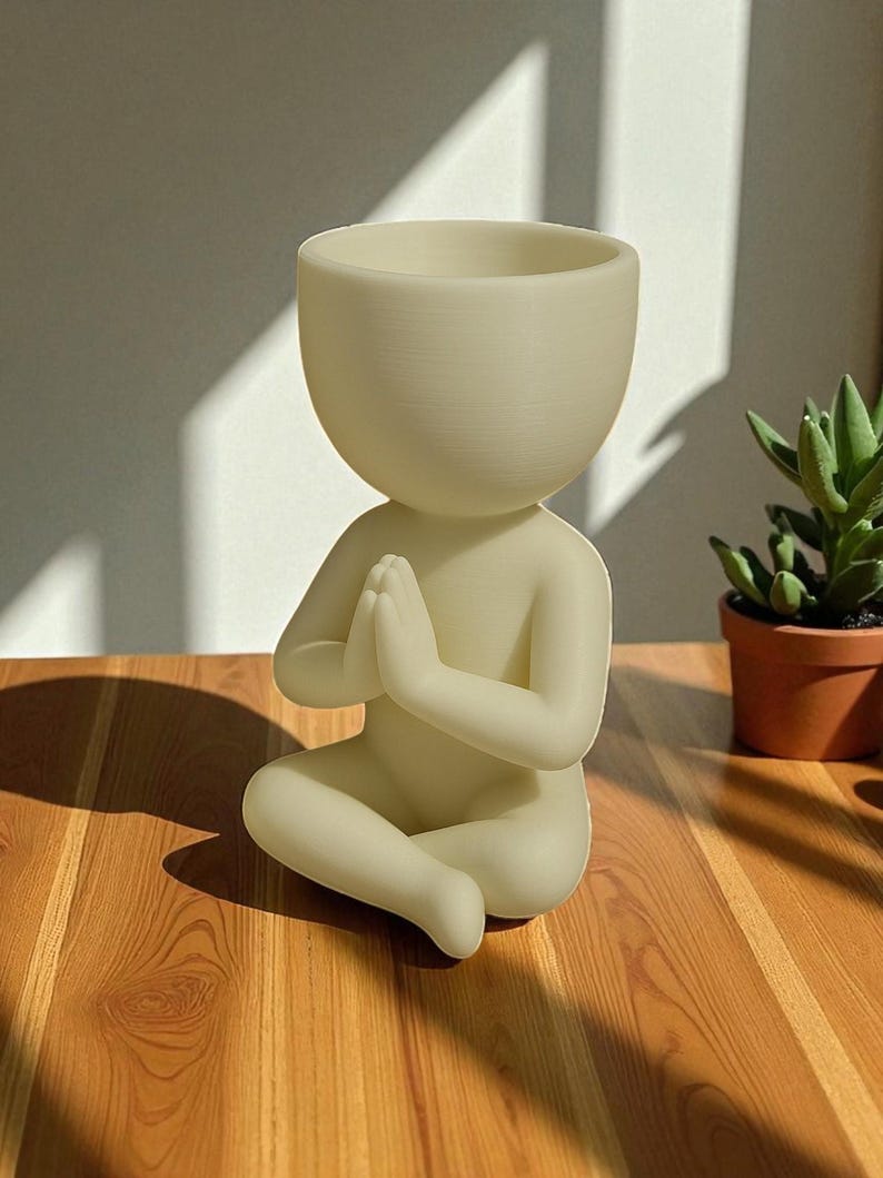 Puede incluir: Una figura crema, sentada con las manos juntas en oraci&oacute;n, dise&ntilde;ada como maceta. La cabeza es un cuenco grande y abierto para plantas. La figura est&aacute; sobre una superficie de madera, con una peque&ntilde;a planta suculenta en maceta al fondo.