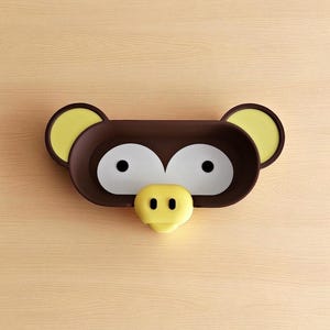 May include: A brown and yellow monkey-shaped wall shelf. The shelf has large white eyes, a yellow nose, and yellow ears. The shelf is mounted on a light brown wooden surface.