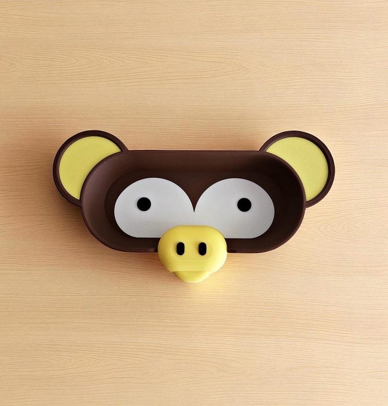 May include: A brown and yellow monkey-shaped wall shelf. The shelf has large white eyes, a yellow nose, and yellow ears. The shelf is mounted on a light brown wooden surface.