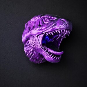 Może przedstawiać: Metalowa fioletowa rzeźba głowy Tyrannosaurus Rex z otwartą paszczą, odsłaniającą ostre zęby i niebieskie wnętrze. Rzeźba jest zamontowana na ciemnoszarej powierzchni, tworząc uderzający kontrast.
