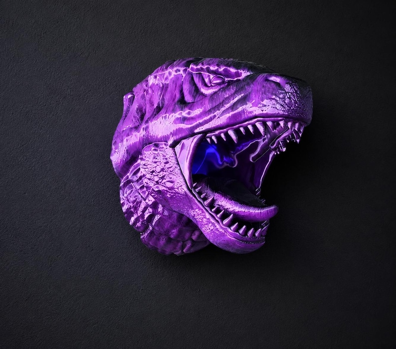 Może przedstawiać: Metalowa fioletowa rzeźba głowy Tyrannosaurus Rex z otwartą paszczą, odsłaniającą ostre zęby i niebieskie wnętrze. Rzeźba jest zamontowana na ciemnoszarej powierzchni, tworząc uderzający kontrast.