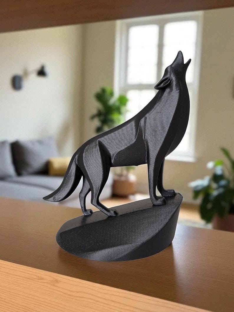May include: A black, geometric wolf sculpture in a howling pose. The wolf is standing on a dark, oval-shaped base. The sculpture is made of a matte material and is set against a blurred background.