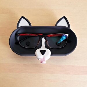 Pode incluir: Um suporte de &oacute;culos preto em forma de gato, com orelhas e nariz brancos. Cont&eacute;m um par de &oacute;culos com arma&ccedil;&atilde;o preta e detalhes vermelhos. A l&iacute;ngua do gato &eacute; rosa.
