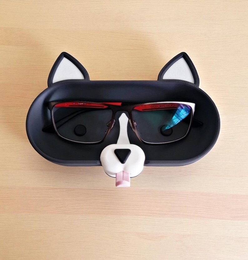 Pode incluir: Um suporte de &oacute;culos preto em forma de gato, com orelhas e nariz brancos. Cont&eacute;m um par de &oacute;culos com arma&ccedil;&atilde;o preta e detalhes vermelhos. A l&iacute;ngua do gato &eacute; rosa.