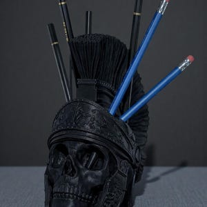 Puede incluir: Un organizador de escritorio negro con forma de calavera con un dise&ntilde;o de casco romano. El organizador contiene l&aacute;pices y pinceles. Los l&aacute;pices son negros y azules con gomas rosas. Los pinceles son negros.